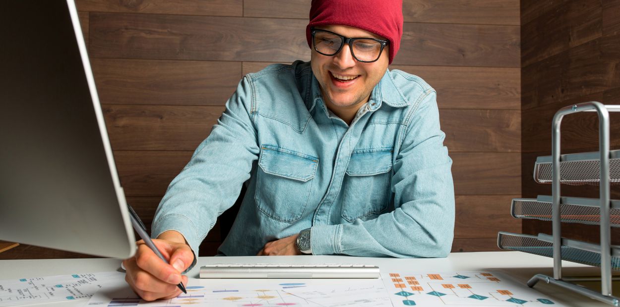 A man is wearing a jean shirt and red hat and sitting at a desk. He's writing on papers.